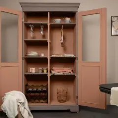 Une armoire relookée aux portes rose corail et intérieur gris, garnie de verres, assiettes, bouteilles et d'une couverture.
