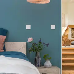 Un lit douillet avec des coussins roses et blancs sur un mur couleur bleu canard. À côté, un escalier en bois mène à un salon cosy.