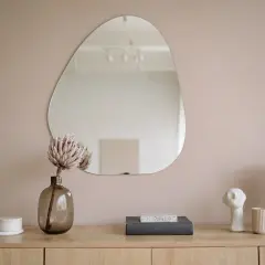 Un miroir aux formes organiques reflète la pièce. Sur le meuble en bois, une fleur séchée, des livres, une lampe en rotin et des objets décoratifs créent une scène apaisante.