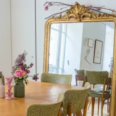 Un grand miroir au cadre doré orné de fleurs et de branches. Une table en bois est dressée avec des vases fleuris, prête à repeindre son cadre doré.