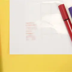 Un cutter rouge et trois feutres (rouge, violet, vert) sont prêts pour un projet créatif. Une **lettre noire** est visible sur le papier de fond.