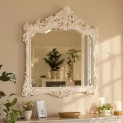 Un miroir ancien en bois, relooké en blanc, orné de volutes et de feuillages, reflète une pièce lumineuse avec des plantes.