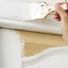 Une main applique de la peinture blanche sur une porte intérieure en bois. Le travail de peinture est en cours, révélant le bois naturel sous la couche blanche.