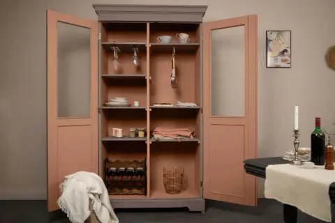 Armoire ancienne en bois vernis relookée, aux portes rose corail et intérieur gris. Elle présente des verres suspendus, des assiettes, des bocaux et une cave à vin.