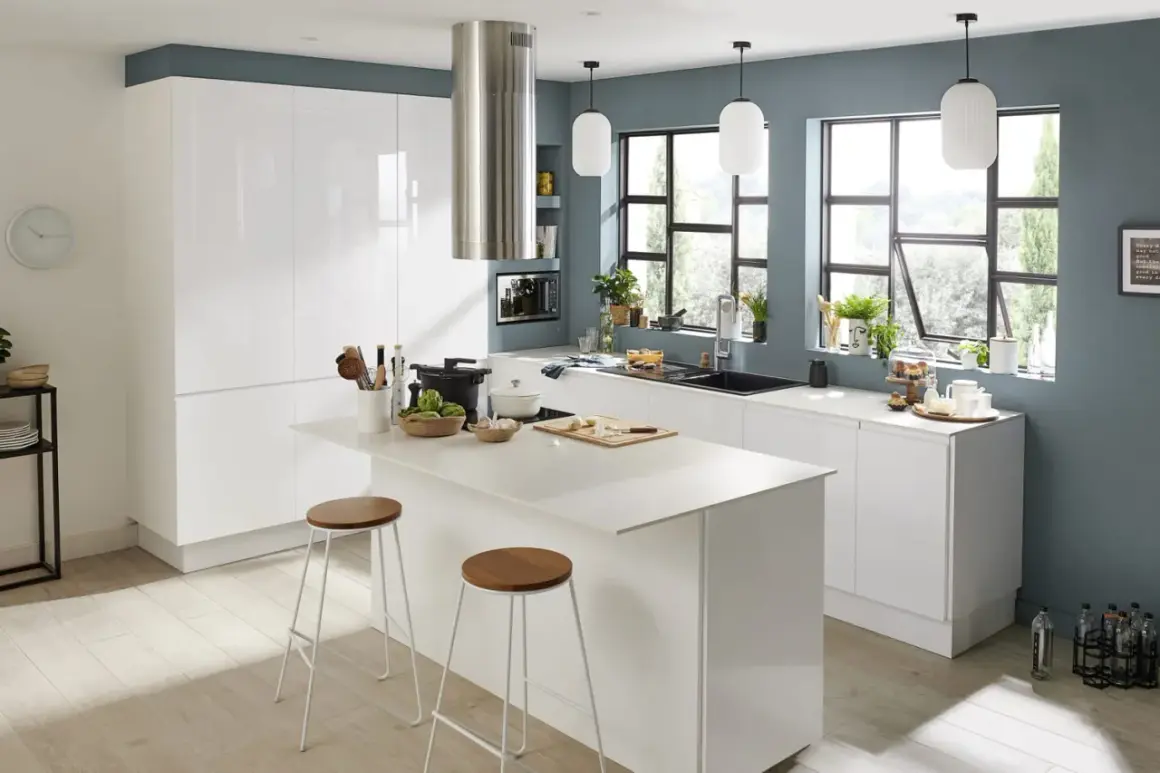 Cuisine moderne aux murs bleu ardoise, avec îlot central blanc, tabourets en bois et métal. Une **peinture acrylique** pourrait capturer l'ambiance lumineuse et épurée.