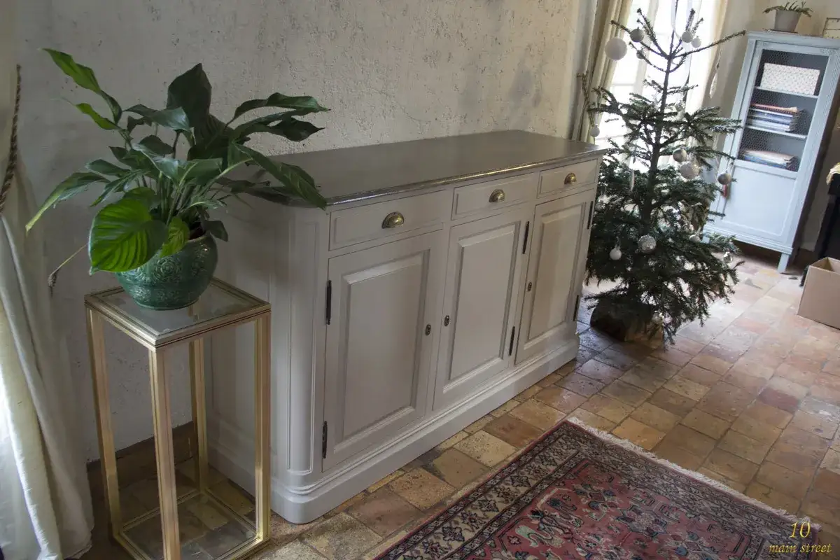 Buffet gris patiné, style ancien, avec un dessus en métal. Une plante verte sur un guéridon doré à gauche, un sapin décoré à droite. Peinture acrylique.